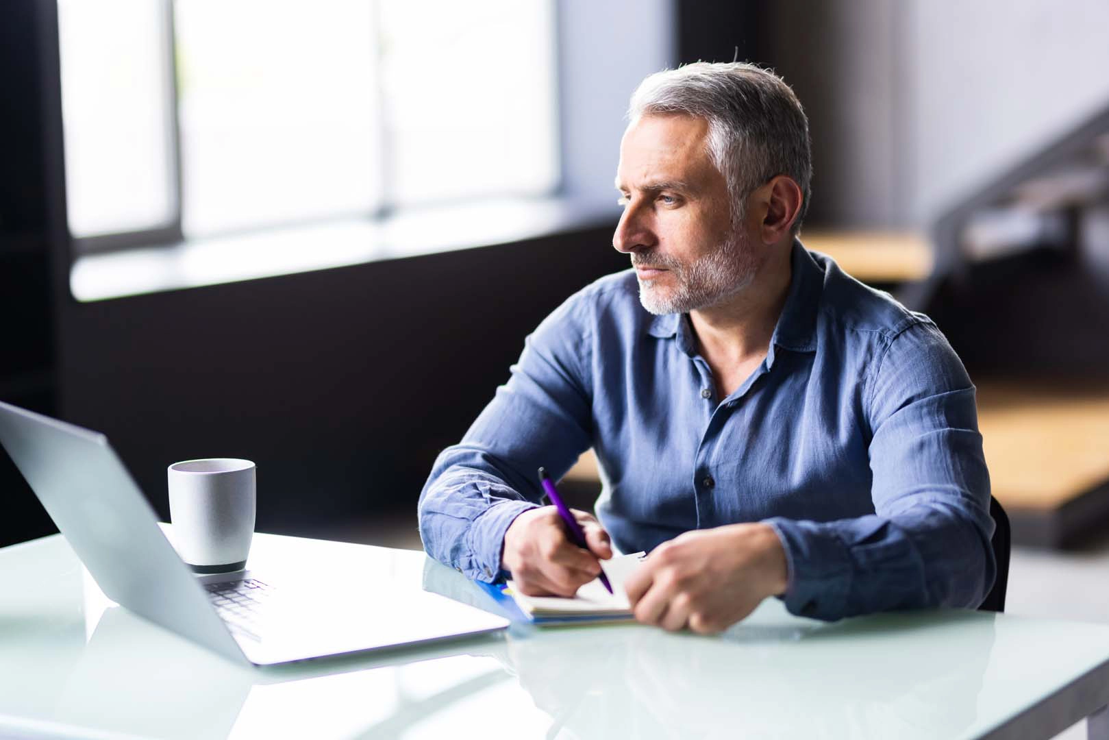 A man at a desk writes on a pad while looking at a laptop screen